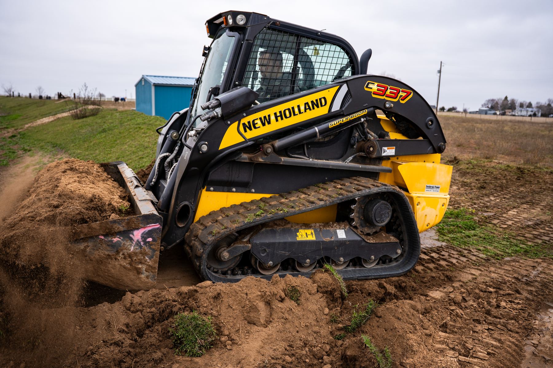 Compact track loader working a hillside near a blue outbuilding.