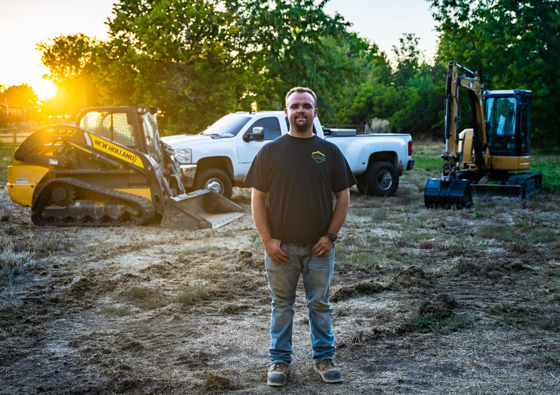 Ollan Stinar standing in a cleared field at sunset with his track loader, truck, and mini excavator behind him.