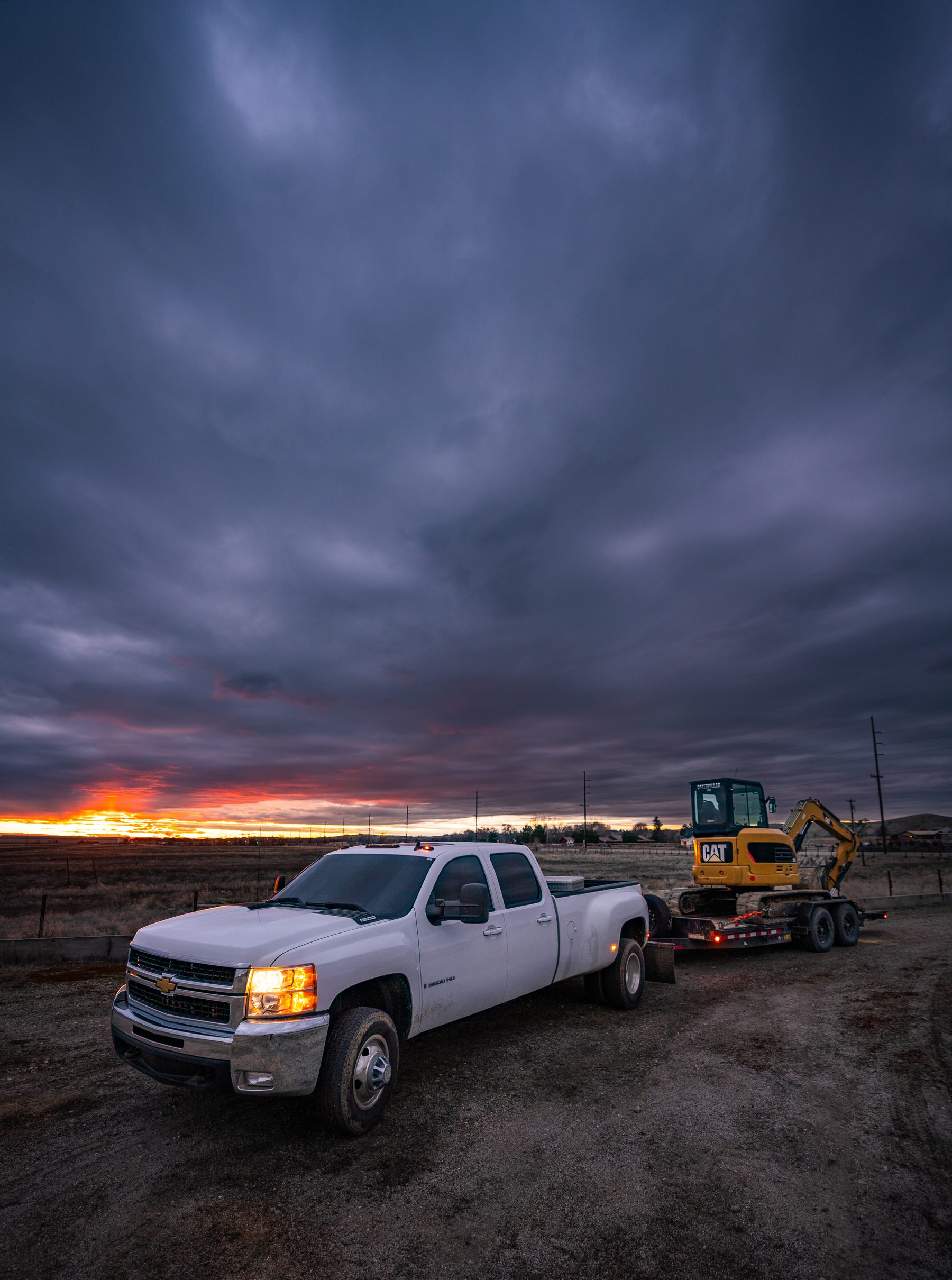 Truck and trailer hauling a mini excavator at dusk.
